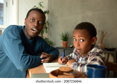 Indoor Picture Of Dark-skinned Male Father Sitting With His Son At Desk In Living-room Doing Homework, Having Shocked Face Expressions, Scared Of Exam Failure, Plate With Cookies And Mug On Table