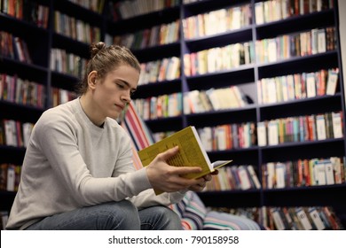 Indoor Picture Of Handsome 18 Year Old Young Man With Hair Bun Sitting At Modern Bookstore, Holding Book In Yellow Cover In His Hands, Having Serious Thoughtful Expression On His Clean Shaven Face