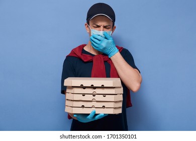 Indoor Picture Of Ill Sneezing Young Courier Covering Mouth With Hand, Wearing Mask And Gloves, Closing Eyes, Being Infected With Coronavirus, Feeling Unwell, Holding Carton Boxes In One Hand.