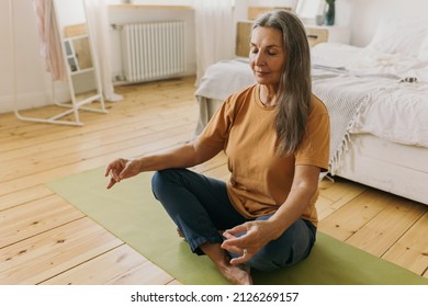 Indoor Picture Of Sporty Mature Female Doing Mudra Sign With Hands, Breathing Deeply, Mediating Keeping Eyes Closed Sitting On Floor On Green Mat Against Bedroom Background With Bed, Window And Mirror