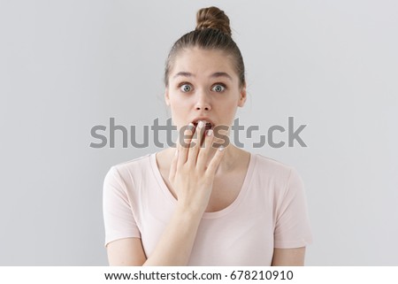 Indoor picture of surprised European female in casual outfit without make up isolated on gray background, looking astonished and deeply shocked, slightly covering round open mouth with hand.