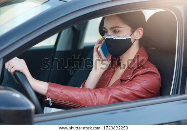 Indoor picture of talkative attractive young\
woman driving car, holding smartphone, having conversation over\
mobile phone, breaking rules, wearing antibacterial mask to protect\
from covid19.