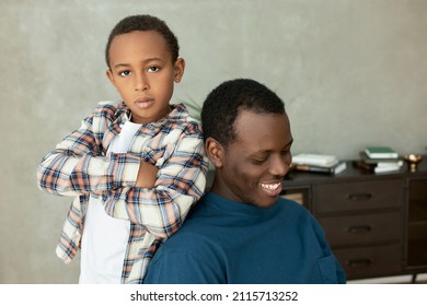 Indoor Picture Of Two Siblings, Little Serious Elementary School Boy With Hands Crossed Leaning Against His 20-years-old Brother Sitting In Chair Against Living-room Interior, Smiling Looking Down