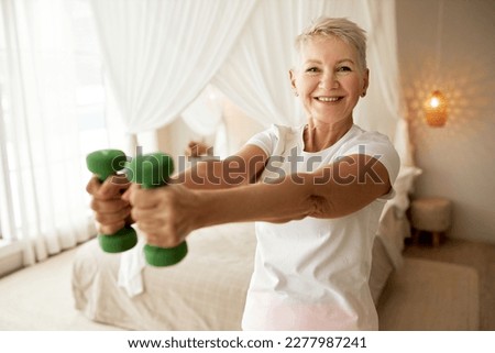 Indoor portrait of 50-years-old woman doing weightlifting exercises at home, holding green small dumbells in outstretched hands, smiling at camera posing against bedroom background