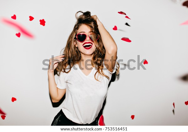 彼女の長い輝く髪に触れる トレンディなtシャツを着た すばらしい白人女性モデルの室内ポートレート 赤い紙吹雪でサングラスをかけた上品な女性が笑う の写真素材 今すぐ編集
