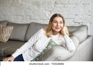 Indoor Portrait Of Attractive Happy 60 Year Old Senior Woman With Pleasant Smile Relaxing On Grey Sofa In Her Modern Apartment Looking In Anticipation While Waiting For Her Children Or Grandchildren