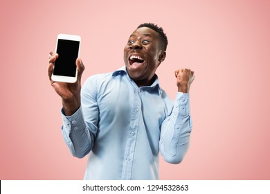 Indoor portrait of attractive young black african man isolated on pink background, holding blank smartphone, smiling at camera, showing screen, feeling happy and surprised. Human emotions, facial