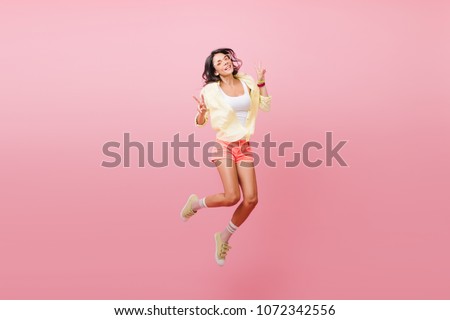 Indoor portrait of beautiful brunette woman with bronze tan fooling around in studio. Slim female model in trendy summer clothes enjoying freedom and smiling.