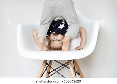 Indoor Portrait Of Beautiful Toddler Dressed In Stylish Pajamas With Stars, Trying To Do Somersault On White Chair In Children's Room. Caucasian Preschool Kid Having Fun And Playing On His Own At Home