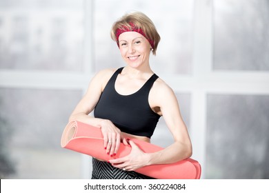 Indoor Portrait Of Cheerful Attractive Fit Senior Woman Posing Holding Red Fitness Mat, Working Out In Sports Club Class, Happy Smiling, Looking At Camera With Friendly Expression