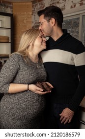 Indoor portrait of a couple in their 30s embracing in the kitchen. The woman is pregnant and is holding a pair of baby shoes. Her husband is kissing her head.