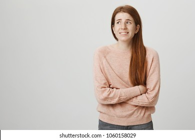 Indoor portrait of cute ginger girl standing with crossed hands on belly, feeling awkward or suffering from pain while looking aside, standing against gray background. Woman has stomachache