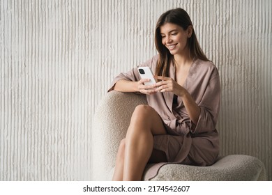 Indoor Portrait Of Cute Lovely Female In Armchair With Smartphone, Wearing Dressing Gown Shopping Online, Watching Stories, Funny Memes On Social Media