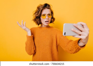 Indoor portrait of disappointed short-haired girl in glasses making selfie in studio. Upset young woman in knitted sweater waving hand during video call, holding smartphone on yellow background. - Powered by Shutterstock