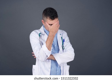 Indoor portrait of doctor man, wearing medical uniform, making facepalm gesture while smiling, standing over gray background amazed with stupid situation.