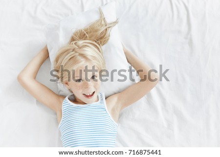 Indoor portrait of emotional blue eyed lovely female child with blonde hair and freckled face having fun in the morning after awakening, feeling excited about new day. Cute little girl lying in bed