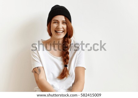 Indoor portrait of fashionable and cheerful young woman with braid wearing stylish black hat and white t-shirt smiling happily while posing in studio. Female fashion, beauty and advertisement concept
