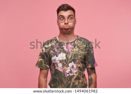 Indoor portrait of funny young handsome tatooed male with glasses grimacing and making faces, posing over pink background with hands down