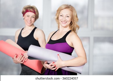 Indoor Portrait Of Group Of Two Cheerful Attractive Fit Senior Women Posing Holding Fitness Mats, Working Out In Sports Club Class, Happy Smiling, Looking At Camera With Friendly Expression