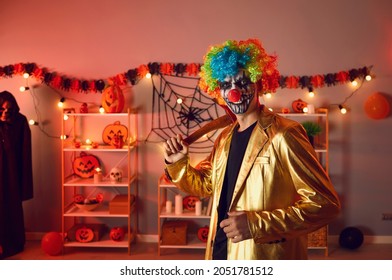 Indoor Portrait Of Guy In Costume Of Spooky Evil Clown At Halloween Party. Adult Man Wearing Gold Suit, Colorful Wig And Fake Red Nose, With Creepy Makeup On Face Holding Axe And Looking At Camera