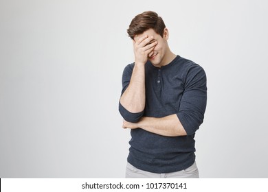 Indoor portrait of handsome man with stylish haircut, wearing casual clothes, making facepalm gesture while smiling, standing over gray background. Guy amazed with stupid situation