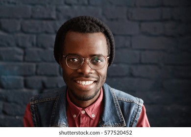 Indoor Portrait Of Happy And Cheerful Young Dark-skinned Male Student Wearing Trendy Clothing Looking At Camera With Joyful Smile While Having Rest In Modern Cafe Interior With Black Brick Walls