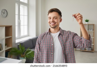 Indoor Portrait Of Happy Male Home Owner Showing Key To His New House. Joyful Handsome Single Young Caucasian Man In Casual Shirt Standing In Living Room, Holding Keys And Smiling. Real Estate Concept