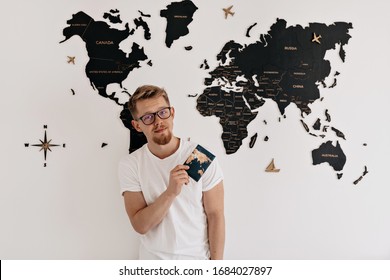 Indoor portrait of happy young european man with passport posing at camera over world map. Preparing for travelling, vacation trip