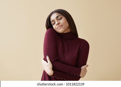 Indoor portrait of positive relaxed young mixed race brunette woman closing eyes with pleasure, keeping arms around herself, enjoying soft fabric of her new maroon cashmere turtleneck sweater