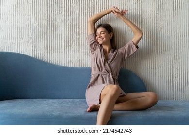 Indoor portrait of relaxed woman in dressing gown having rest on blue couch stretching hands up, smiling with closed eyes on sunday morning with copy space on the right side of picture