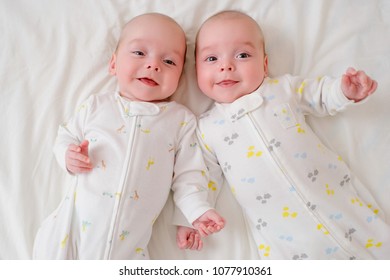 Indoor Portrait Of Two Cute Twin Babies Wearing One Piece Pajamas, Lying Down In Bed And Having Fun. High Angle Shot, View From Above. Friendship And Brotherhood Concept