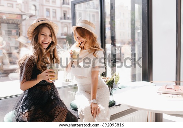 Indoor portrait of two stylish sisters relaxing in cafe after shopping. Smiling long-haired girl with glass of juice in hand posing in front of window next to friend in trendy vintage hat.