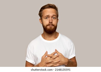 Indoor portrait of young attractive red-haired man with beard looking pitiful with sad face expression and scared eyes, isolated on grey background, standing with clasped hands and interlocked fingers
