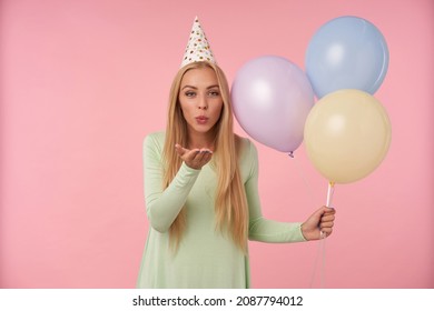 Indoor Portrait Of Young Blonde Female, Wears Green Dress, Party Hat Holding Balloons And Send Air Kiss Into Camera.posing Over Pink Background