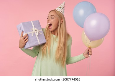 Indoor Portrait Of Young Blonde Female, Wears Green Dress, Party Hat Holding Balloons And Big Gift. Posing Over Pink Background