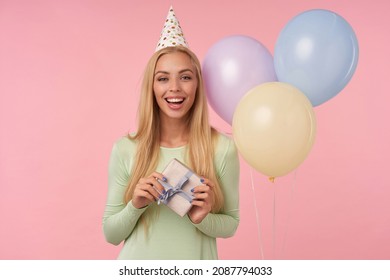 Indoor Portrait Of Young Blonde Female, Wears Green Dress, Party Hat Holding Small Gift Box Smiles Broadly And Feels Excited, Posing Over Pink Background