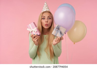 Indoor Portrait Of Young Blonde Female, Wears Green Dress, Party Hat Holding Small Gift, Feels Happy And Smiles Broadly. Posing Over Pink Background