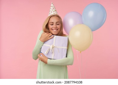 Indoor Portrait Of Young Blonde Female, Wears Green Dress, Party Hat Hug Her Big Gift Box And Feels Happy And Excited Posing Over Pink Background