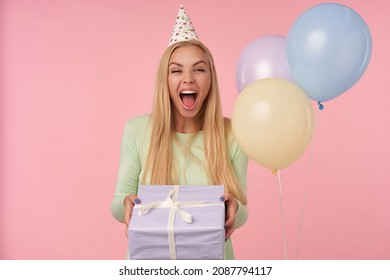 Indoor Portrait Of Young Blonde Female, Wears Green Dress, Party Hat Posing Holding Her Gift Box With Surprised Facial Expression. Over Pink Background