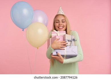 Indoor Portrait Of Young Blonde Female, Wears Green Dress, Party Hat Holding Few Gifts, Smiles Broadly And Feels Satisfied, Posing Over Pink Background