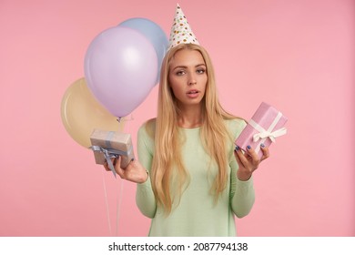 Indoor Portrait Of Young Blonde Female, Wears Green Dress, Party Hat Posing Over Pink Feels Confused While Choose The Gift. Background