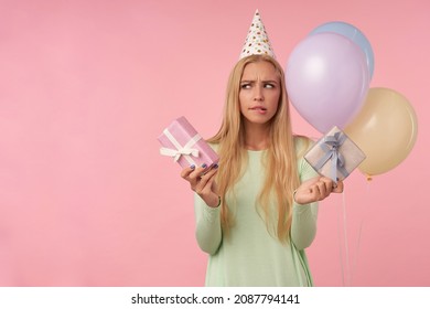 Indoor Portrait Of Young Blonde Female, Wears Green Dress, Party Hat Posing Over Pink Feels Confused While Choose The Gift. Background