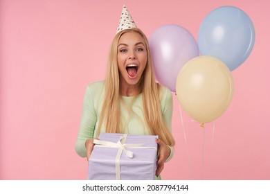 Indoor Portrait Of Young Blonde Female, Wears Green Dress, Party Hat Posing Holding Her Gift Box With Surprised Facial Expression. Over Pink Background