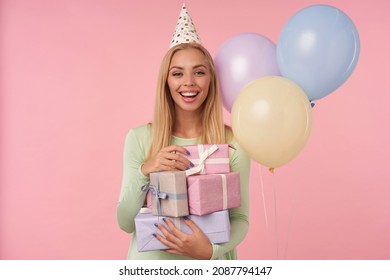 Indoor Portrait Of Young Blonde Female, Wears Green Dress, Party Hat Holding Few Gifts And Smiles Broadly. Posing Over Pink Background