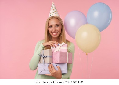 Indoor Portrait Of Young Blonde Female, Wears Green Dress, Party Hat Holding Few Gifts, Smiles Broadly And Feels Satisfied, Posing Over Pink Background