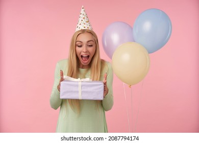 Indoor Portrait Of Young Blonde Female, Wears Green Dress, Party Hat Posing Holding Her Gift Box With Surprised Facial Expression. Over Pink Background