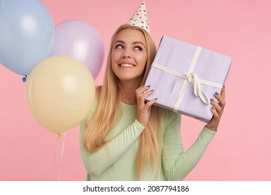 Indoor Portrait Of Young Blonde Female, Wears Green Dress, Party Hat Posing Holding Her Gift Box With Surprised Facial Expression. Over Pink Background