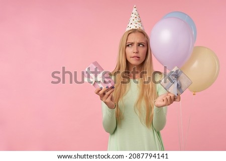 indoor portrait of young blonde female, wears green dress, party hat posing over pink feels confused while choose the gift. background