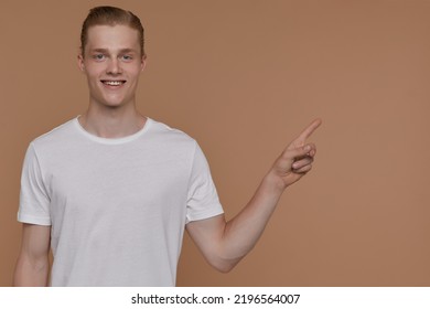 Indoor Portrait Of Young Blonde Man Posing Over Beige Background Point With A Finger Aside At Copy Space With Excited Facial Expression