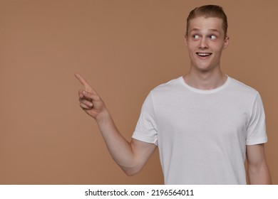 Indoor Portrait Of Young Blonde Man Posing Over Beige Background Point With A Finger Aside At Copy Space With Excited Facial Expression